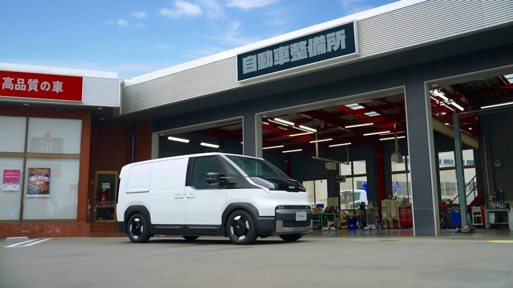 Right side view of the Kia PV5 parked in front of a service repair shop.